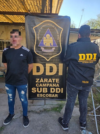 Hombre pixelado en camiseta negra y jeans rotos junto a un agente policial de espaldas, frente a un cartel de la DDI de la Provincia de Buenos Aires