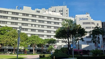 Un gran edificio blanco de varios pisos con muchas ventanas uniformes y el nombre 'Jackson' en azul. Hay árboles verdes y palmeras en primer plano bajo un cielo azul
