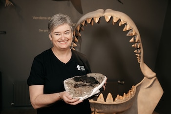 Mujer de cabello gris sonriendo sostiene un fósil. Detrás se ve una réplica de una mandíbula de tiburón. El fondo es oscuro y tiene texto en una pared