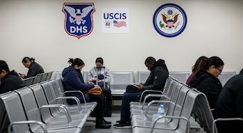 Una sala de espera con bancos metálicos y varias personas sentadas, algunas con maletas, bajo los logos del DHS y USCIS en la pared blanca.