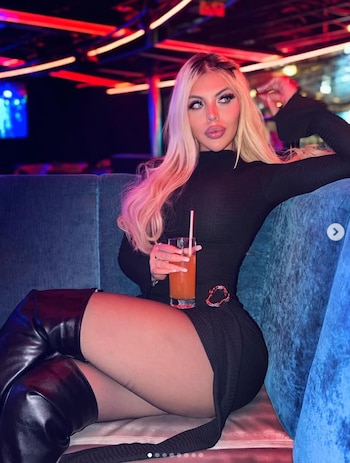 Una mujer rubia con cabello largo y un vestido negro ajustado está sentada en un sofá de terciopelo azul en un club, sosteniendo un trago