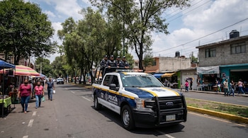 Camioneta de policía blanca y azul con oficiales de pie en la parte trasera, circulando por una calle concurrida en Tláhuac, con árboles y edificios.