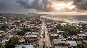 Vista aérea de un poblado junto al mar bajo la lluvia, con vehículos que circulan por calles anegadas y nubes oscuras en el cielo.