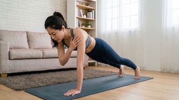 Mujer con camiseta deportiva gris y leggings azules en posición de plancha, tocándose el hombro con una mano sobre una esterilla de yoga azul.