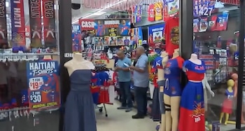 Interior de tienda con maniquíes exhibiendo ropa azul y roja. Un cartel anuncia camisetas haitianas en oferta. Dos hombres y más mercadería visible
