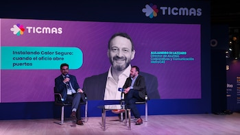 Dos hombres sentados en sillas, uno con micrófono, en un escenario. Detrás, una pantalla grande con el logo de Ticmas, un título y la foto de Alejandro Di Lazzaro