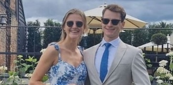Hombre y mujer sonrientes con gafas de sol. Hombre con traje gris y corbata azul. Mujer con vestido azul y blanco. Fondo exterior con árboles y sombrillas