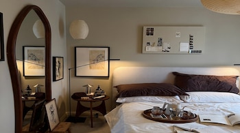Una habitación con una cama con sábanas blancas y almohadas marrones, un espejo de marco de madera, una bandeja con tetera y tazas, y arte en las paredes.