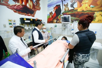 En una sala de hospital, el Ministro de Salud, un médico y una enfermera observan a un paciente en una camilla, con equipos médicos y un mural de El Rey León de fondo