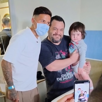 Un hombre con mascarilla azul y tatuajes posa junto a un adulto que sostiene a un niño, mientras una mano los enfoca con un teléfono móvil