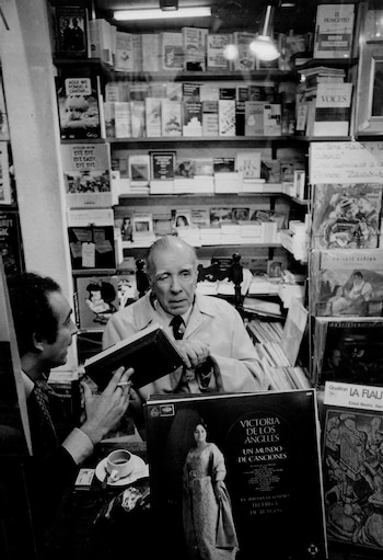 Hombre mayor con gafas leyendo un libro en una librería abarrotada de estanterías con libros y discos de vinilo. Una segunda persona se ve parcialmente a la izquierda