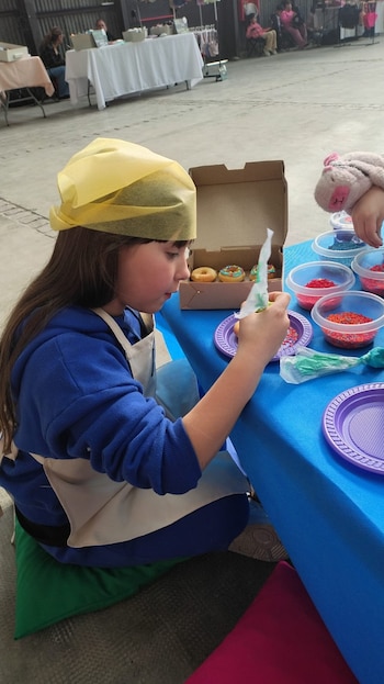 Una niña con pañuelo amarillo y delantal aplica cobertura a una rosquilla sobre una mesa azul con recipientes de chispas