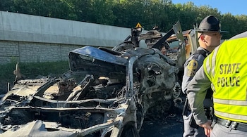 Restos carbonizados de un automóvil y parte de un camión visible en una carretera con un muro de contención. Dos oficiales de policía, uno con un chaleco "STATE POLICE", observan la escena