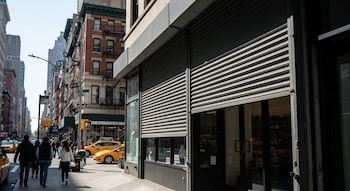 Vista frontal de una calle de Nueva York con una tienda que tiene una persiana metálica gris parcialmente bajada. Personas y taxis amarillos transitan por la acera.