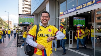 Hombre con camiseta de Colombia sostiene bandera. Personas con indumentaria deportiva salen de edificio. Pantallas muestran partido Colombia vs Suiza Mundial 2026.