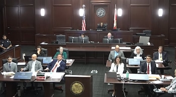 Un tribunal con un juez en el estrado, banderas de Estados Unidos y Florida, sellos oficiales. Abogados y personal sentados en mesas con laptops, un oficial uniformado