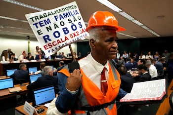 El legislador y pastor Sargento Isidorio sostiene un cartel en apoyo al fin de la semana laboral de seis días en Brasil, que dice en portugués: "Los trabajadores tienen familias y no son robots. ¡Ya debemos implementar la jornada laboral 5x2!", durante una sesión especial del comité que analizaba la propuesta en la Cámara de Diputados, el miércoles 27 de mayo de 2026, en Brasilia, Brasil (AP Foto/Eraldo Peres)