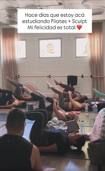 Varias personas, mayormente mujeres, realizan ejercicios de piso con las piernas en alto en una clase de Pilates o Sculpt en un estudio bien iluminado, con una barra de ballet visible