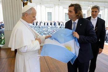 El Papa Francisco, vestido de blanco, mira sonriendo a un hombre en traje azul que sostiene una bandera argentina. Otro hombre de esmoquin negro está detrás
