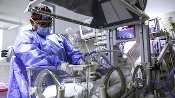 Un médico con bata azul y mascarilla trabaja en una incubadora transparente rodeada de monitores de control.