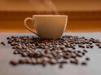 Una taza blanca humeante se encuentra sobre una superficie oscura, rodeada de granos de café tostados; el fondo es de color marrón y está desenfocado