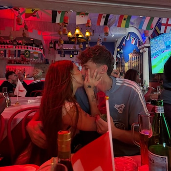 Una pareja se besa; la mujer tiene cabello largo pelirrojo y el hombre viste camiseta azul y sostiene una rosa roja. Al fondo, banderas de países y una bandera suiza