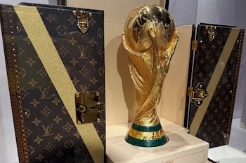 Un trofeo dorado de la Copa Mundial de la FIFA con base verde, flanqueado por dos cofres abiertos con el monograma de Louis Vuitton y franjas doradas