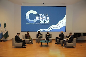 Seis personas sentadas en sillas, frente a una pantalla con el texto 'Conveniencia 2026'. Hay dos banderas institucionales y un piano de cola