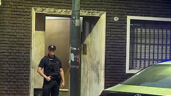 Oficial de policía con chaleco táctico de pie frente a la entrada de un edificio de ladrillos oscuros por la noche, con un coche patrulla visible