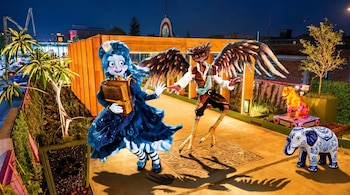 Personaje femenino azul con un libro y personaje alado junto a esculturas de un perro y un elefante sobre una terraza nocturna iluminada.