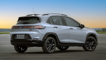 Vista trasera y lateral de un Chevrolet Sonic gris claro, un B-SUV coupé, estacionado en un pavimento oscuro con un cielo azul y naranja al fondo