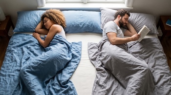 Vista cenital de una mujer que duerme bajo una manta azul y un hombre que lee un libro bajo una manta gris en una cama.