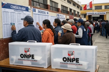 Vista frontal de votantes peruanos revisando paneles electorales en un colegio, con ánforas de la ONPE en primer plano y una bandera de Perú al fondo.