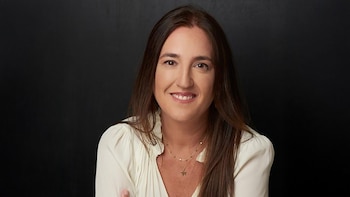 Retrato frontal de una mujer sonriente con cabello castaño largo, luciendo una blusa color crema y collares, sobre un fondo oscuro