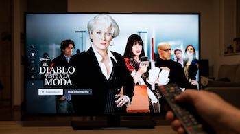 Una televisión muestra el póster de la película "El Diablo Viste a la Moda" con el logo de Disney+. En primer plano, una mano sostiene un control remoto apuntando a la pantalla.