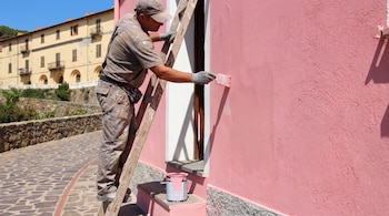 Un hombre subido a una escalera de madera aplica pintura de color rosa en una pared exterior junto a un bote de pintura.