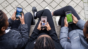 Vista cenital de tres personas sentadas en un banco, cada una con un teléfono inteligente. Las pantallas muestran un paisaje, una persona bailando y un partido de fútbol.