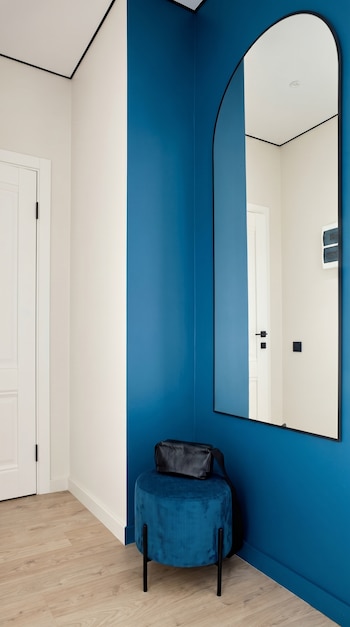Un recibidor moderno con una pared azul cerúleo, un espejo arqueado con marco negro, un taburete de terciopelo azul y una bolsa negra.