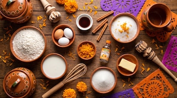 Recipientes de barro con harina, leche y azúcar sobre una mesa de madera adornada con flores de cempasúchil y papel picado.