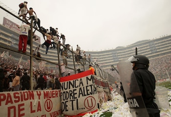 Multitud de hinchas escalan una valla en estadio de fútbol. Un agente policial con casco y escudo en primer plano. El suelo tiene papel picado