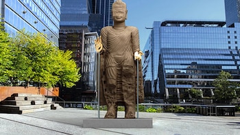 Estatua grande de Buda de color marrón sin rostro y con manos doradas, de pie en un parque urbano con edificios modernos de cristal y árboles verdes al fondo