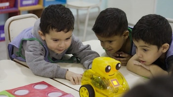 Tres niños con delantales escolares observan un robot de juguete amarillo con ruedas sobre una mesa
