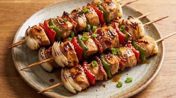 Imagen de primer plano de cuatro brochettes de pollo, pimientos rojos y verdes, cebolla y panceta, con salsa teriyaki, sésamo y verdeo, sobre un plato.