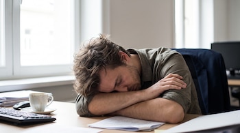 Un hombre duerme con la cabeza apoyada en sus brazos sobre un escritorio con un teclado, un ratón, una taza de café, documentos y una ventana.