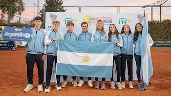 Ocho jóvenes, cinco hombres y tres mujeres, posan sonrientes en una cancha de tenis de tierra batida, sosteniendo una bandera argentina y un cartel azul de "Argentina"