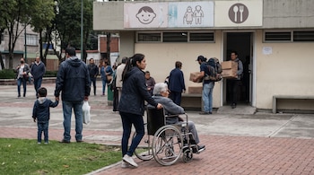 Grupo de personas en plaza pública. Mujer empuja silla de ruedas con adulto. Hombre lleva niño de la mano. Otro hombre entrega cajas. Edificio con letreros de servicios comunitarios.
