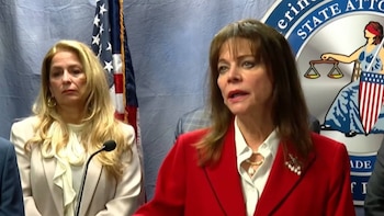 Dos mujeres en un entorno oficial; una con blazer rojo y la otra con blazer claro. Al fondo, la bandera de Estados Unidos y un sello de oficina