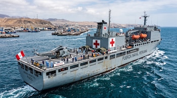 Un buque gris con la bandera de Perú, cruces rojas, un helicóptero en cubierta y personal con uniforme azul cerca de la costa