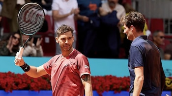 Dos tenistas masculinos en una cancha de arcilla roja; uno, Guido Andreozzi, con camiseta roja, levanta su raqueta; el otro, de espaldas, con camiseta azul