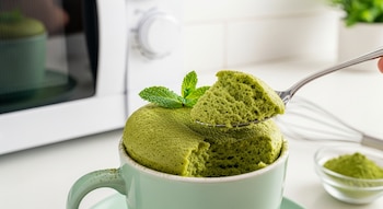 Una mano levanta con una cuchara una porción de bizcocho de té verde recién hecho en una taza verde. Un microondas blanco se ve al fondo.